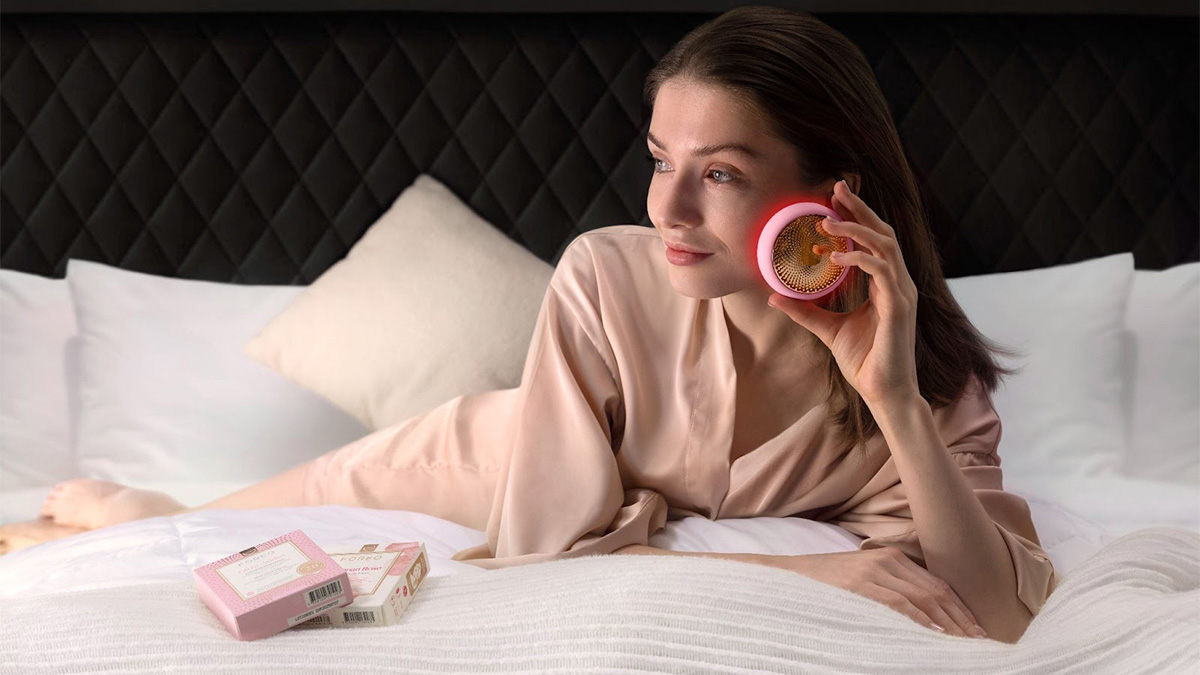 A young woman is lying on her stomach on a comfortable large bed with many pillows. She is wearing a dusty pink robe and using the pink UFO 3 deep facial hydration device